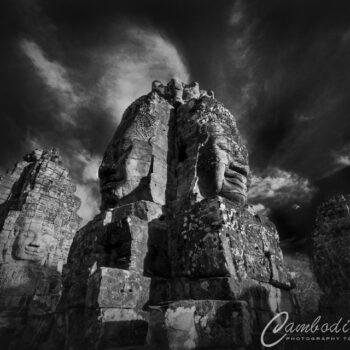 Dramatic fine-art black and white photography of the giant carved stone faces on the towers of Bayon Temple in Angkor, Cambodia, shot from a low angle against a dark, cloudy sky.