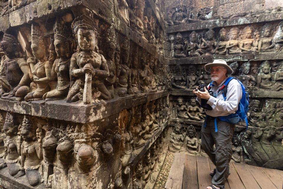 An Angkor iconic photography tour client wearing a sun hat and backpack holds his camera while exploring the deeply carved stone relief walls inside the hidden trench of the Terrace of the Leper King.