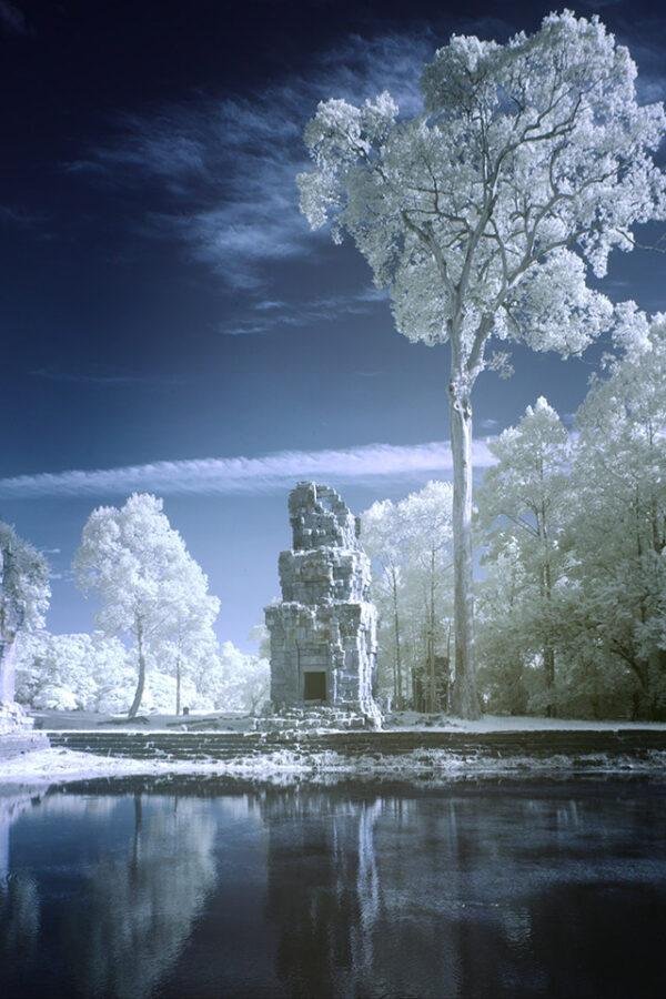 False color infrared photograph of Prasat Suor Prat temple tower in Angkor Thom Cambodia, featuring glowing white trees and dark blue skies reflected in a calm pond.