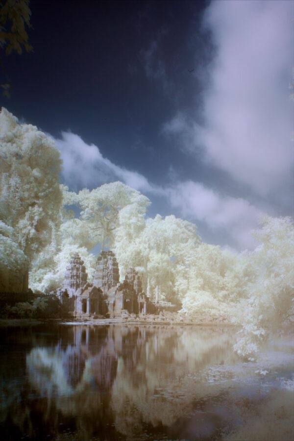 Fine art color infrared photography showing the monumental stone entrance gate towers of Preah Khan temple in Angkor, Cambodia, perfectly reflected on the calm water moat amid glowing white jungle foliage under a dark sky.