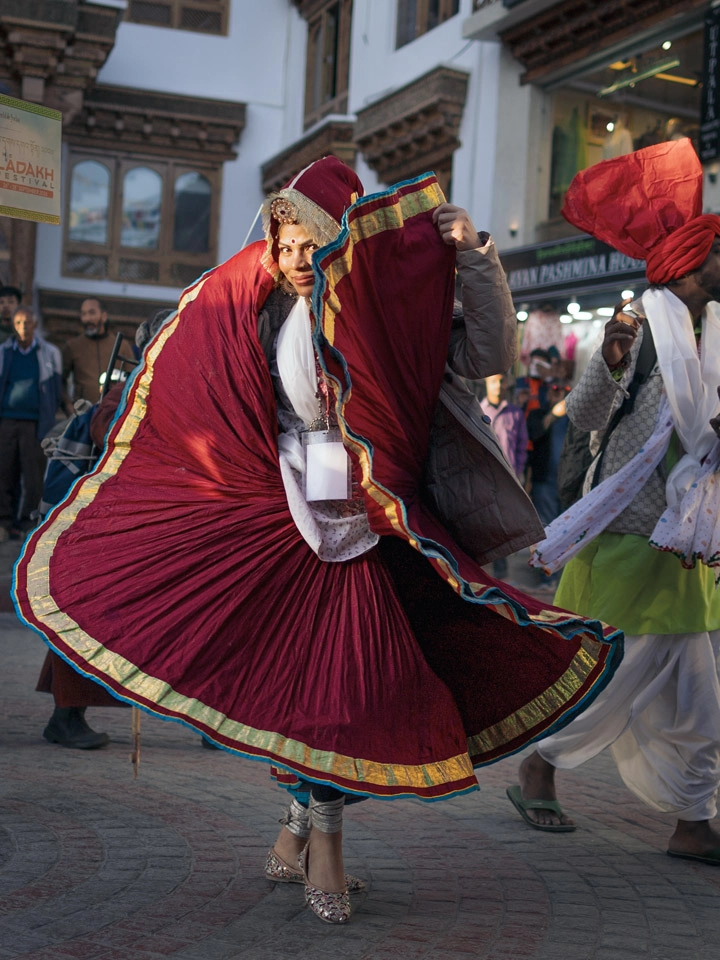 A side-profile view of a dancer spinning dynamically in a wide, pleated dark red dress with gold trim during a festive street performance in Ladakh.