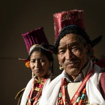portrait of an elderly Ladakhi man and woman smiling warmly while wearing traditional tall, embroidered maroon headwear and coral necklaces.