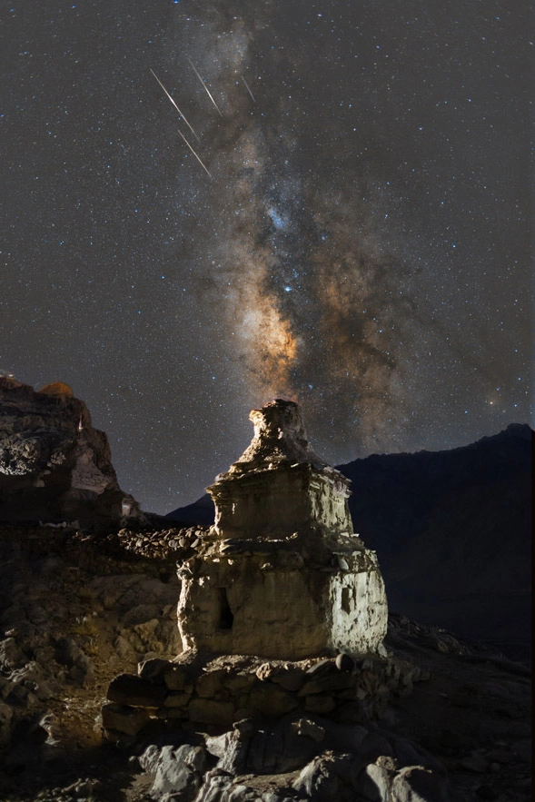An ancient stone Buddhist chorten stupa illuminated at night, with the vibrant Milky Way galaxy and multiple shooting star meteor streaks visible in the dark sky above.